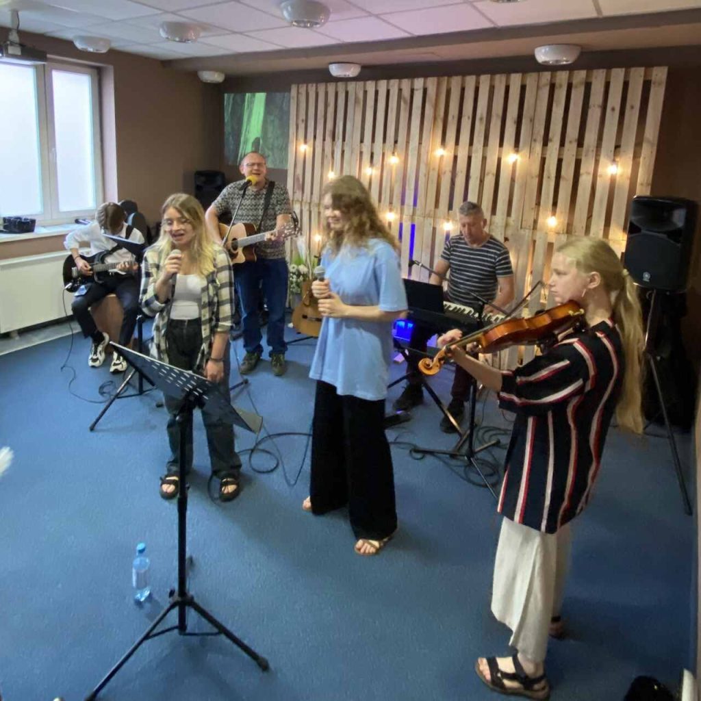 Zespół muzyczny podczas występu w sali, z wokalistkami, gitarzystami, klawiszowcem i skrzypaczką, na tle drewnianej ścianki z oświetleniem.