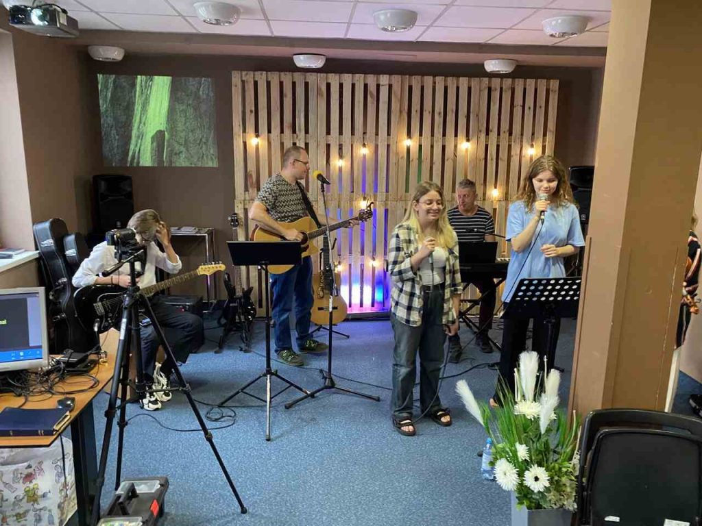 Zespół muzyczny podczas próby lub występu, z dwoma wokalistkami, gitarzystą, klawiszowcem i dekoracyjną drewnianą ścianką z oświetleniem w tle.