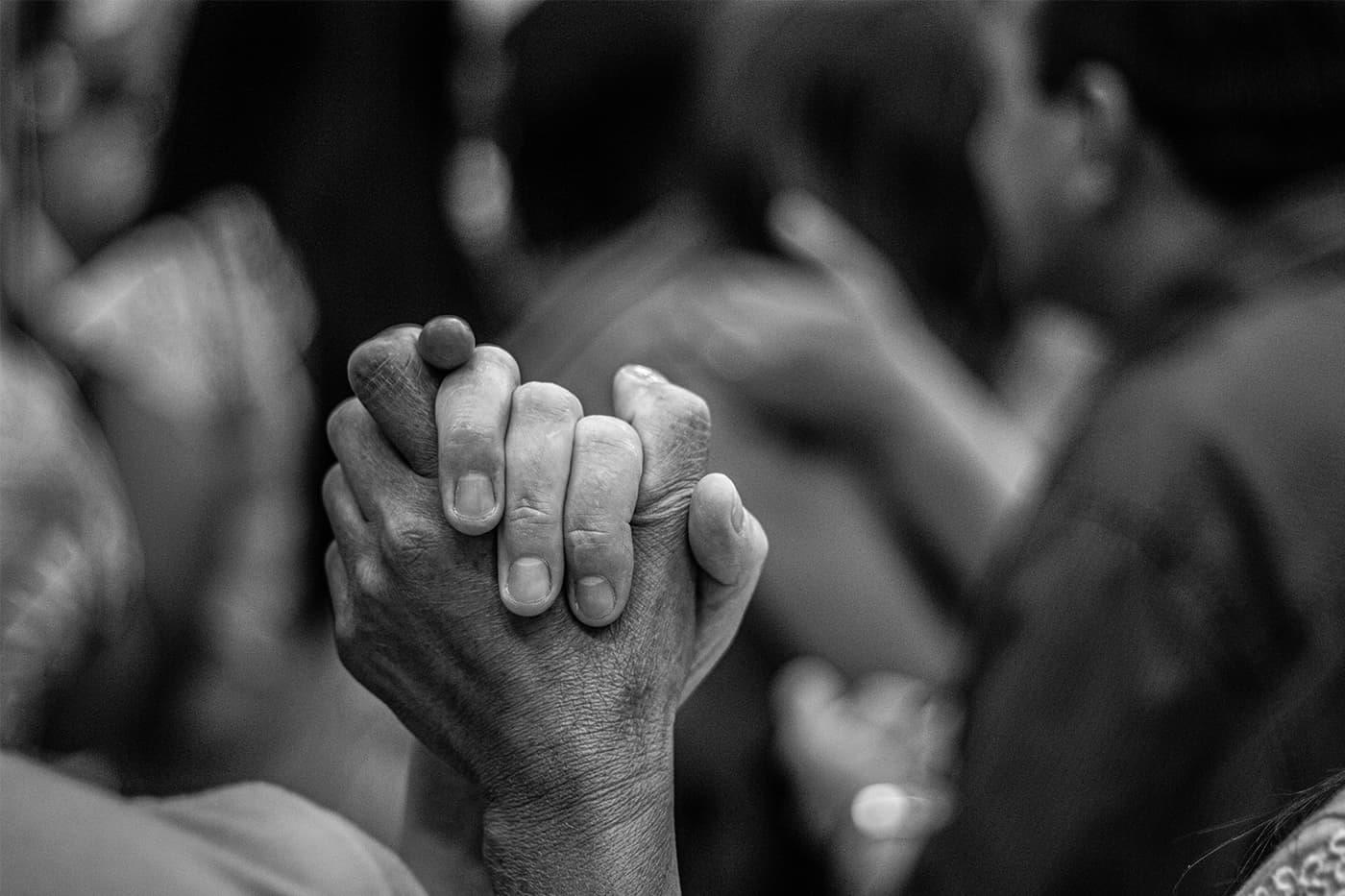Czarno-białe zdjęcie dwóch osób trzymających się za ręce w geście wsparcia i jedności, w tle rozmyte postacie.