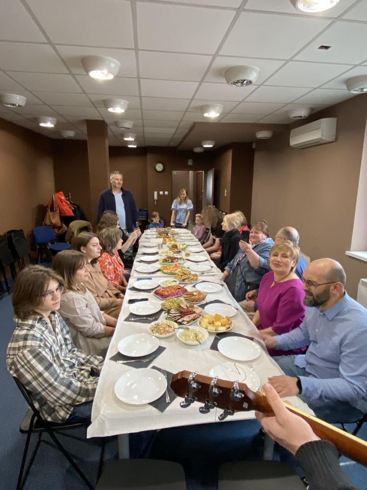 Grupa ludzi siedzących przy długim stole zastawionym jedzeniem podczas spotkania towarzyskiego w sali z brązowymi ścianami i sufitem z lampami.