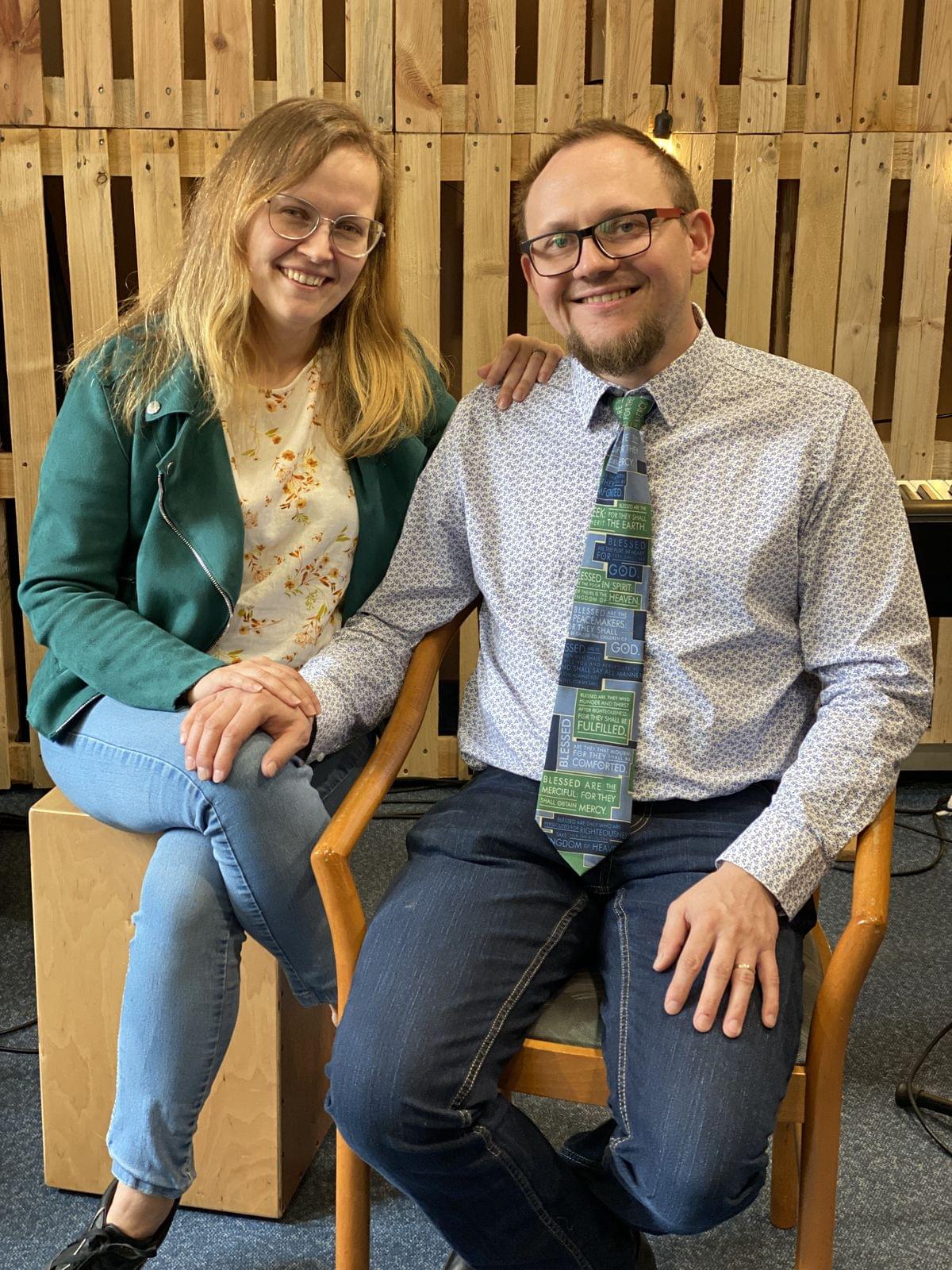 Uśmiechnięta para siedząca razem, trzymająca się za ręce, na tle drewnianej ścianki dekoracyjnej.