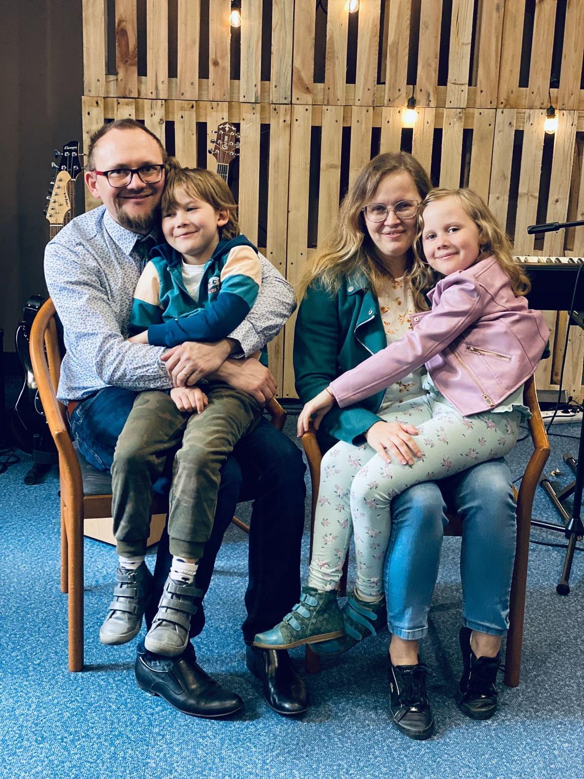 Rodzina siedząca na krzesłach, uśmiechnięci rodzice trzymający dwójkę dzieci na kolanach, na tle drewnianej ścianki dekoracyjnej w jasnej sali.